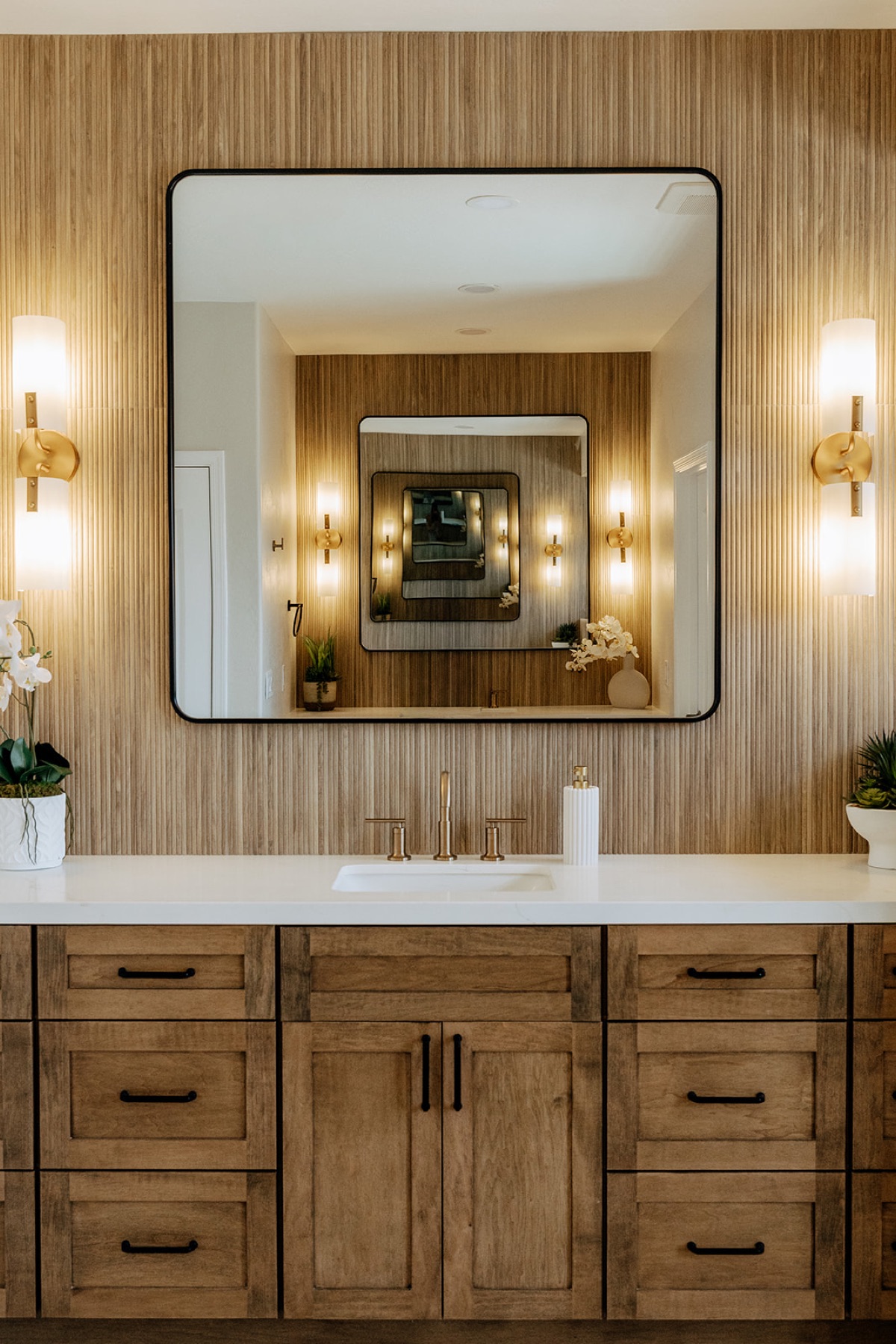 New primary bathroom vanity with a custom stained wood cabinet, reeded tile accent wall, black-framed mirror, and brass fixtures